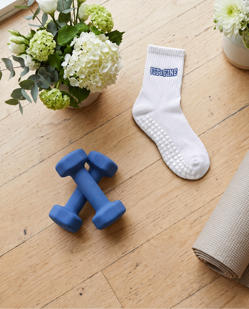 The Fit & Fine grip sock set, styled on a light wood floor with a rolled yoga mat, blue dumbbells, and two flower arrangements.