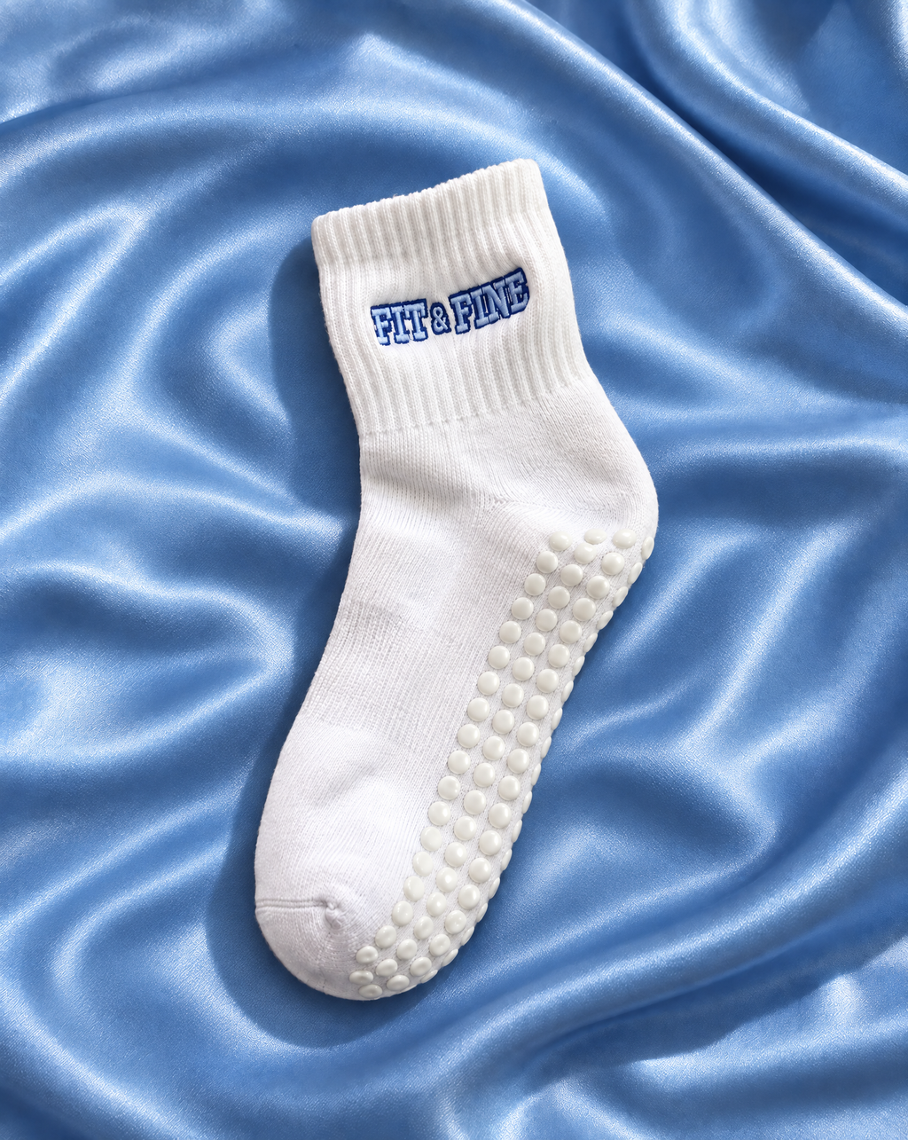 A single white pilates grip sock with 'Fit & Fine' embroidered in blue on the cuff, displayed on a shiny light blue satin fabric background.
