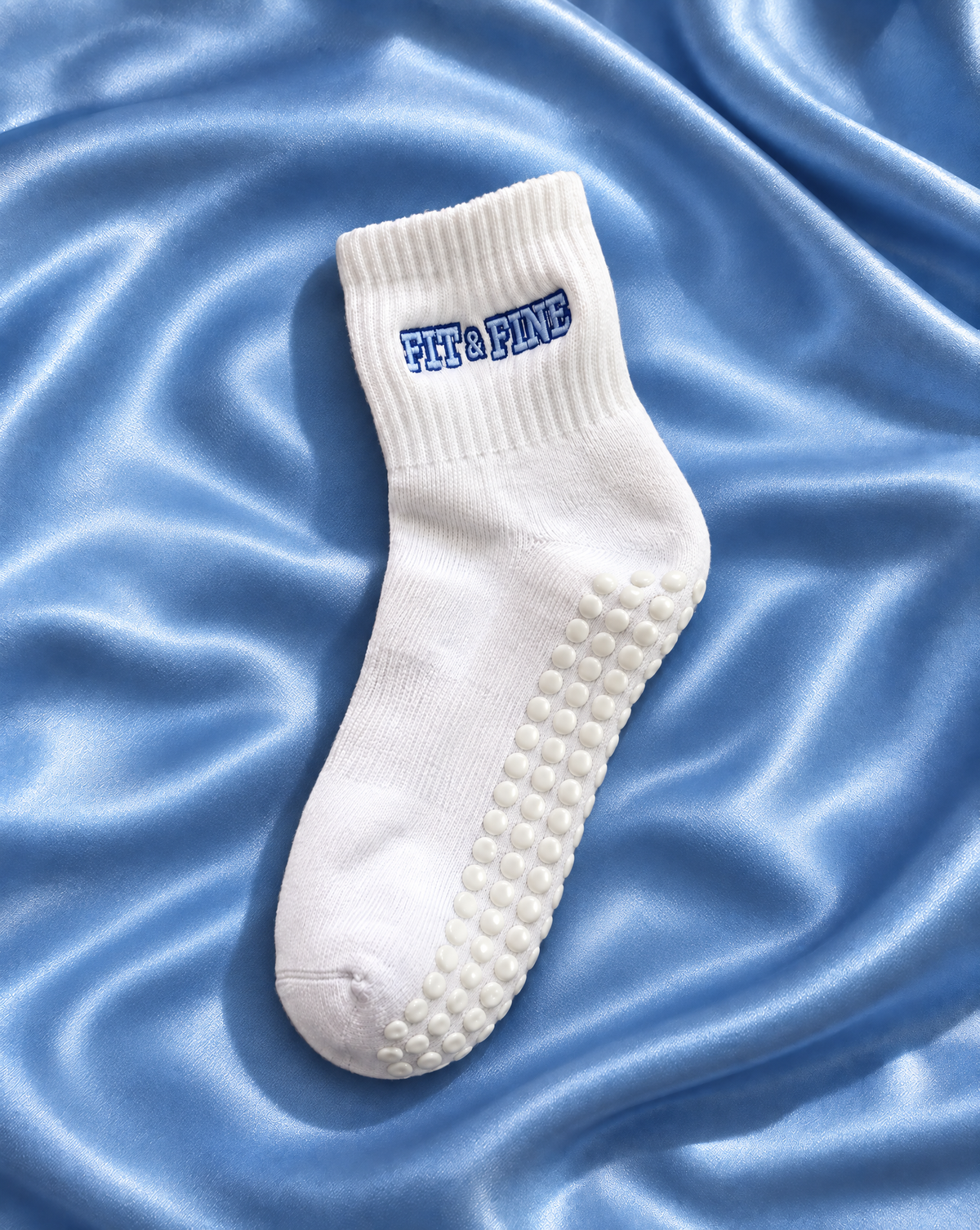 A single white pilates grip sock with 'Fit & Fine' embroidered in blue on the cuff, displayed on a shiny light blue satin fabric background.