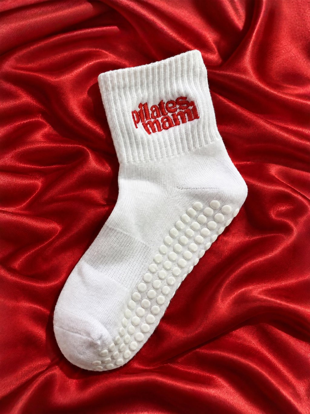 A single white crew sock with red embroidered 'pilates march' text on the cuff and a raised grip pattern on the sole, laid on a shiny red satin background.