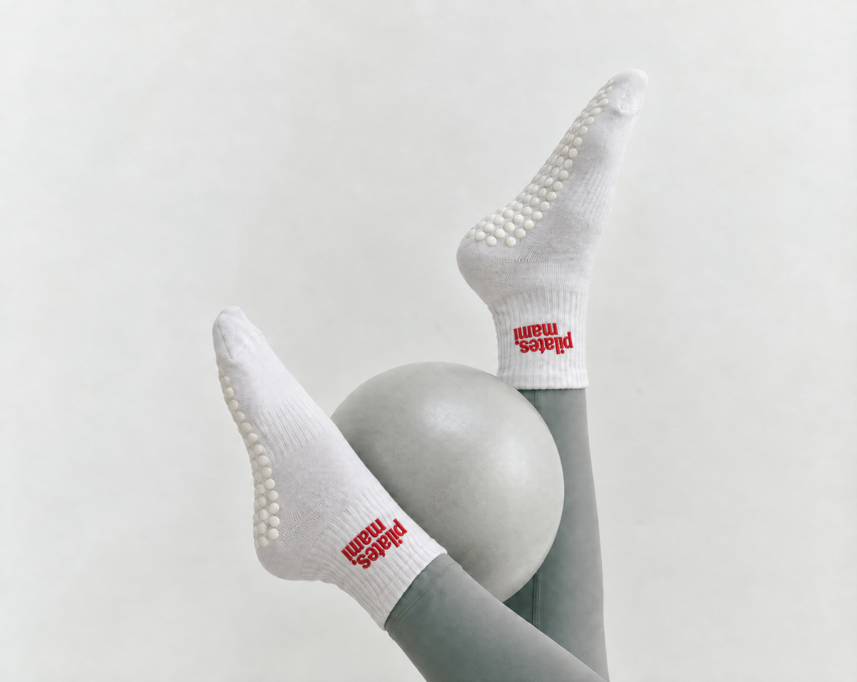 White grip socks with red 'Pilates Mami' detail, worn on a person's legs positioned around a light gray pilates ball against a plain light background.