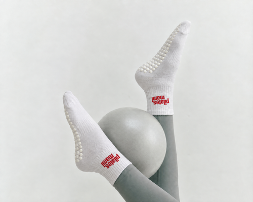 White grip socks with red 'Pilates Mami' detail, worn on a person's legs positioned around a light gray pilates ball against a plain light background.